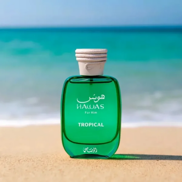 Green perfume bottle labeled 'Hawas' on a sandy beach with ocean in the background